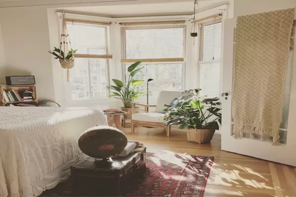 Sunlit bedroom with white bedding, large windows, green houseplants, a wooden chair, and a patterned rug on a hardwood floor.