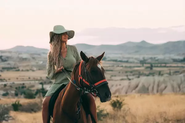 Woman in a green dress and wide-brimmed hat riding a brown horse in a vast, open landscape with mountains in the distance.