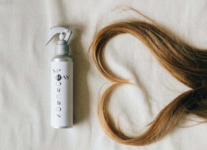 Hair restoration spray bottle next to long healthy blonde hair arranged on a neutral fabric background