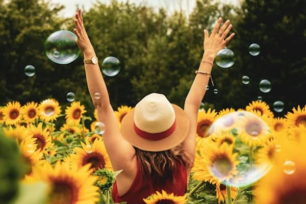 A woman wearing a sunhat and red sleeveless top raises her arms joyfully in a vibrant sunflower field surrounded by floating soap bubbles.