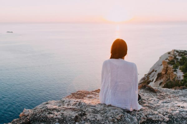 Woman sitting on a cliff at sunset, reflecting quietly over the ocean.